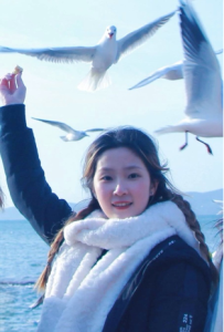 Yihan in a dark coat and fluffy white scarf standing by the sea with one arm raised as seagulls fly overhead