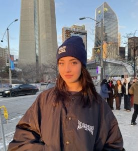 A woman in a black coat and a blue hat standing in front of the CN tower with other people and buildings in the background.